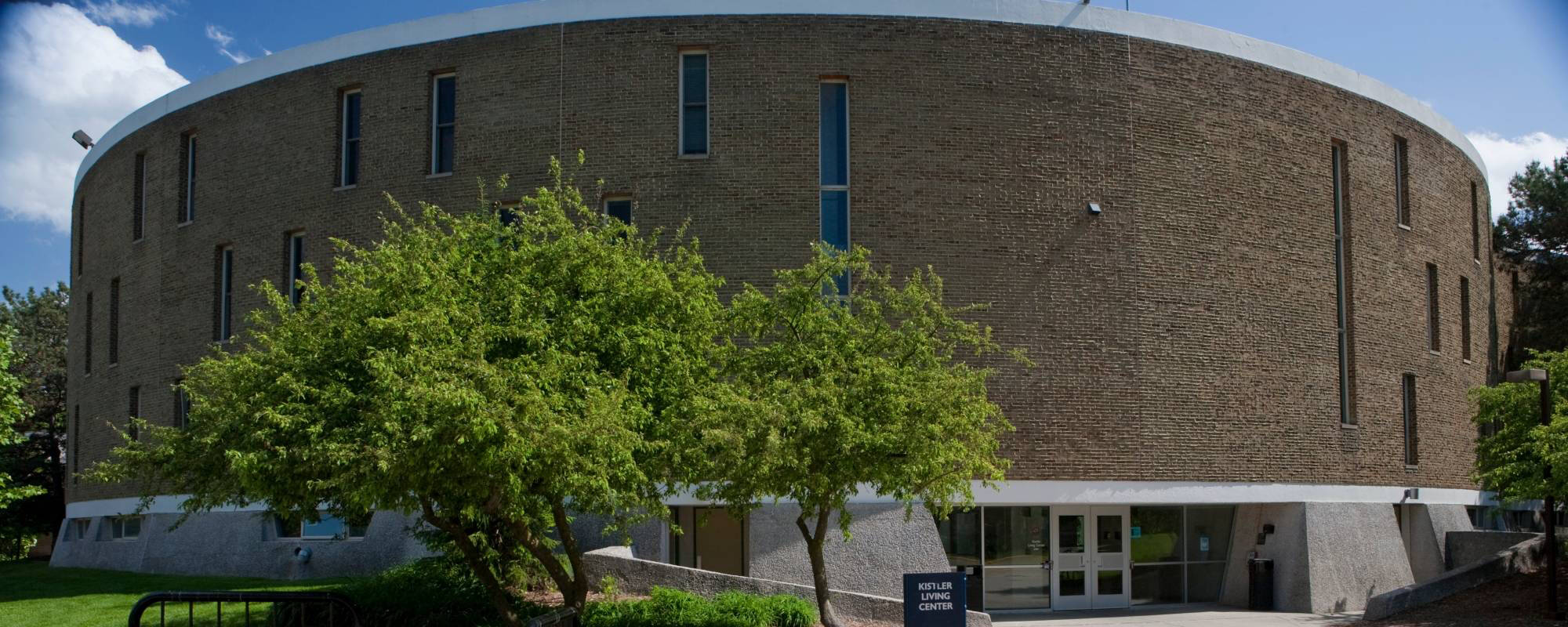 Circular brick building, Kistler Living Center, under a blue sky, with tall narrow windows and green trees in front. The entrance has glass doors and a small sign.